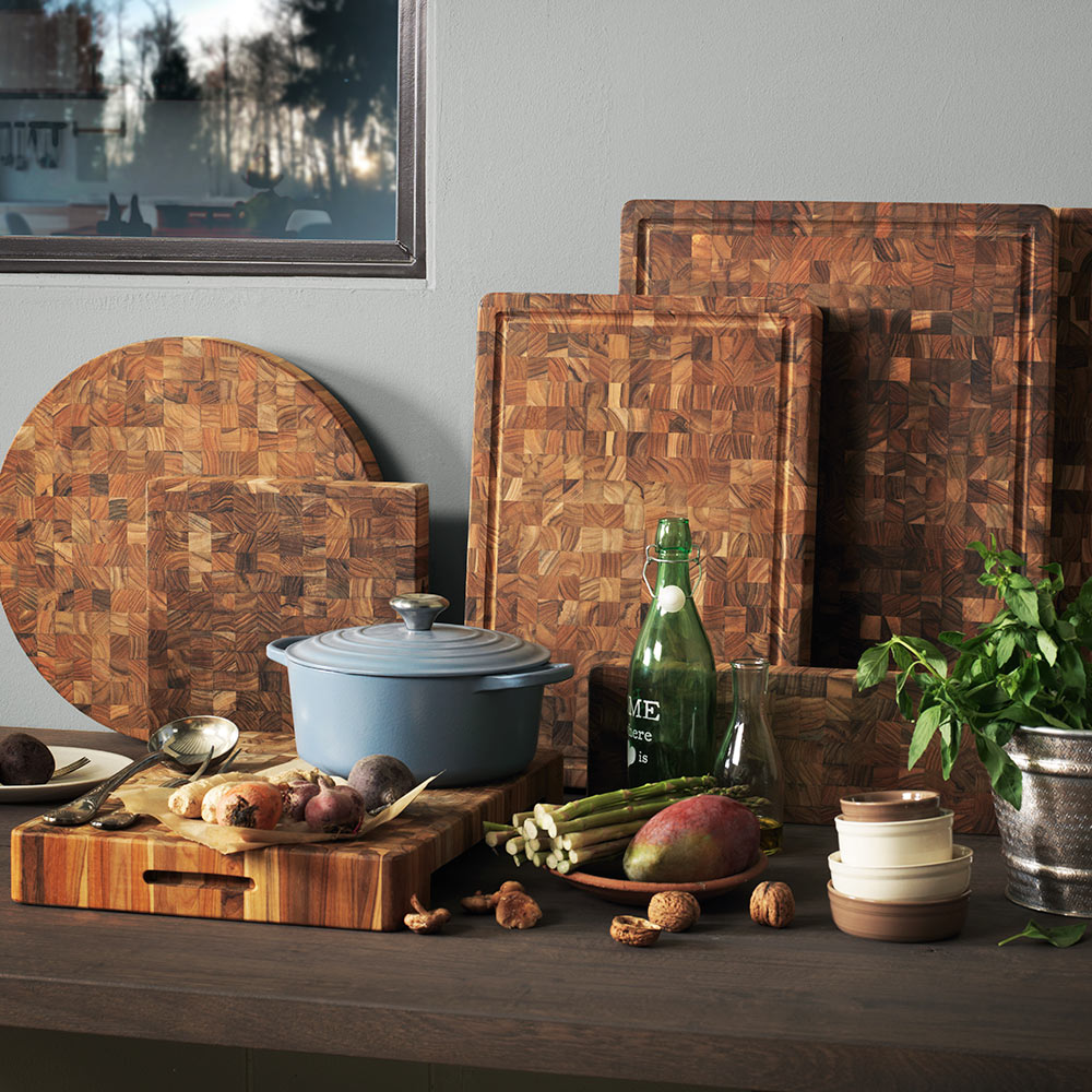 Teak end grain butcher blocks and a round serving board arranged with fresh vegetables and a blue Dutch oven.