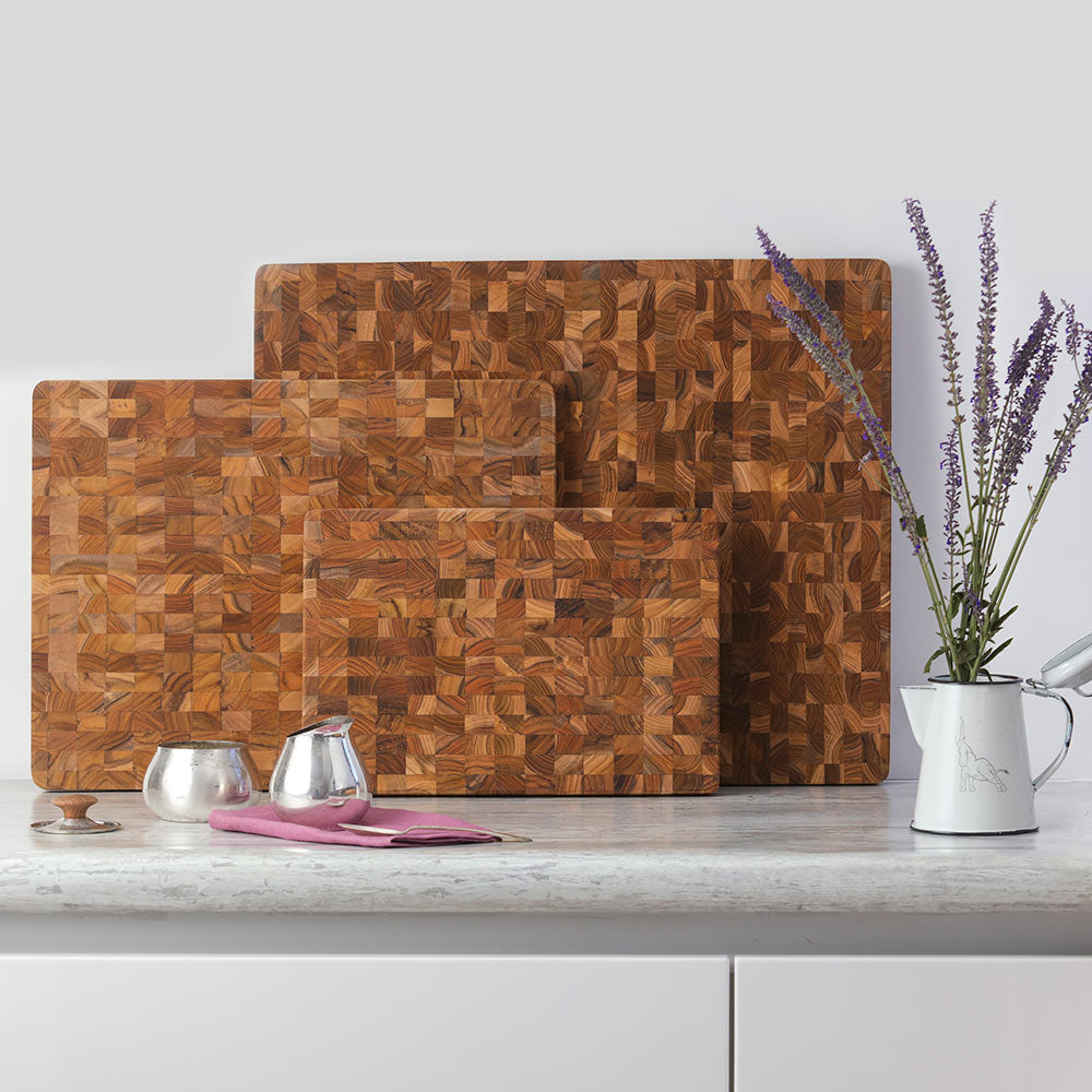 Three thin end grain teak wood cutting boards of varying sizes displayed on a bright marble kitchen counter.