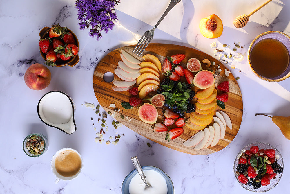Elegant large oval teakwood board displaying colorful sliced fruits on a white marble background.